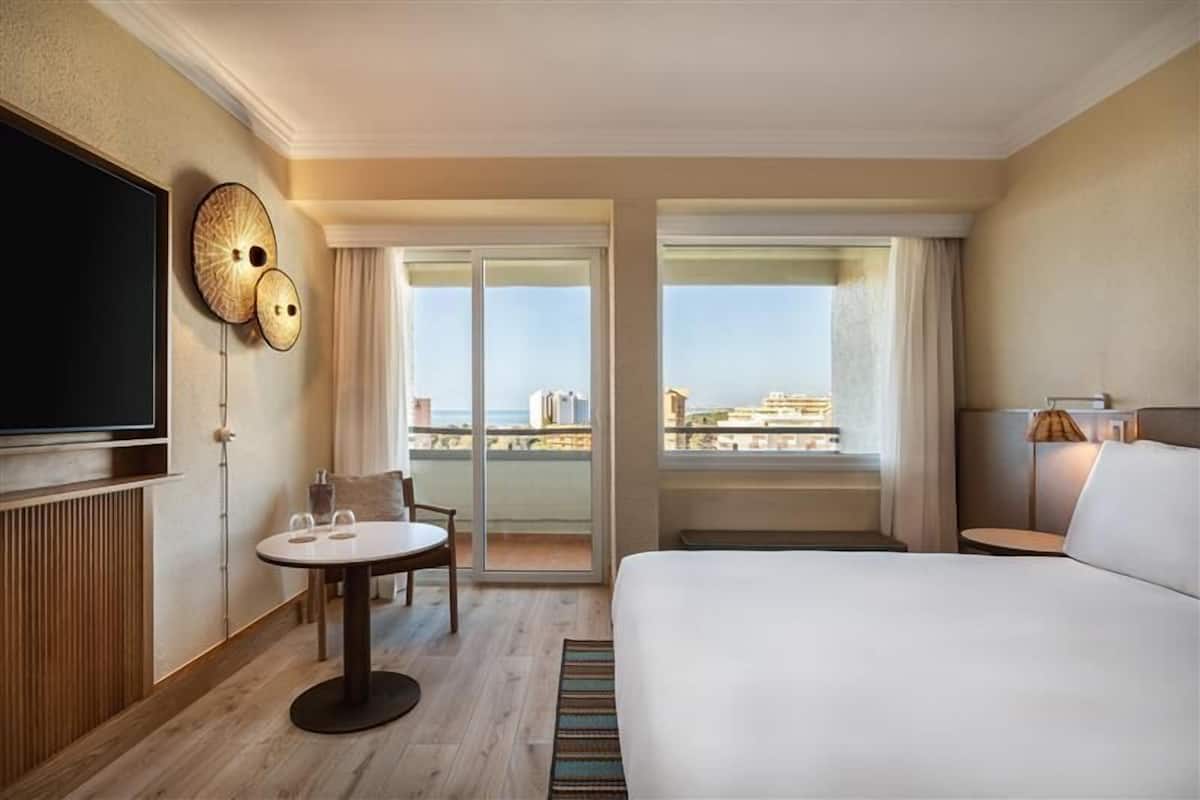 A hotel room with a large bed, a TV, a dining table, and a view of the cityscape through the balcony.