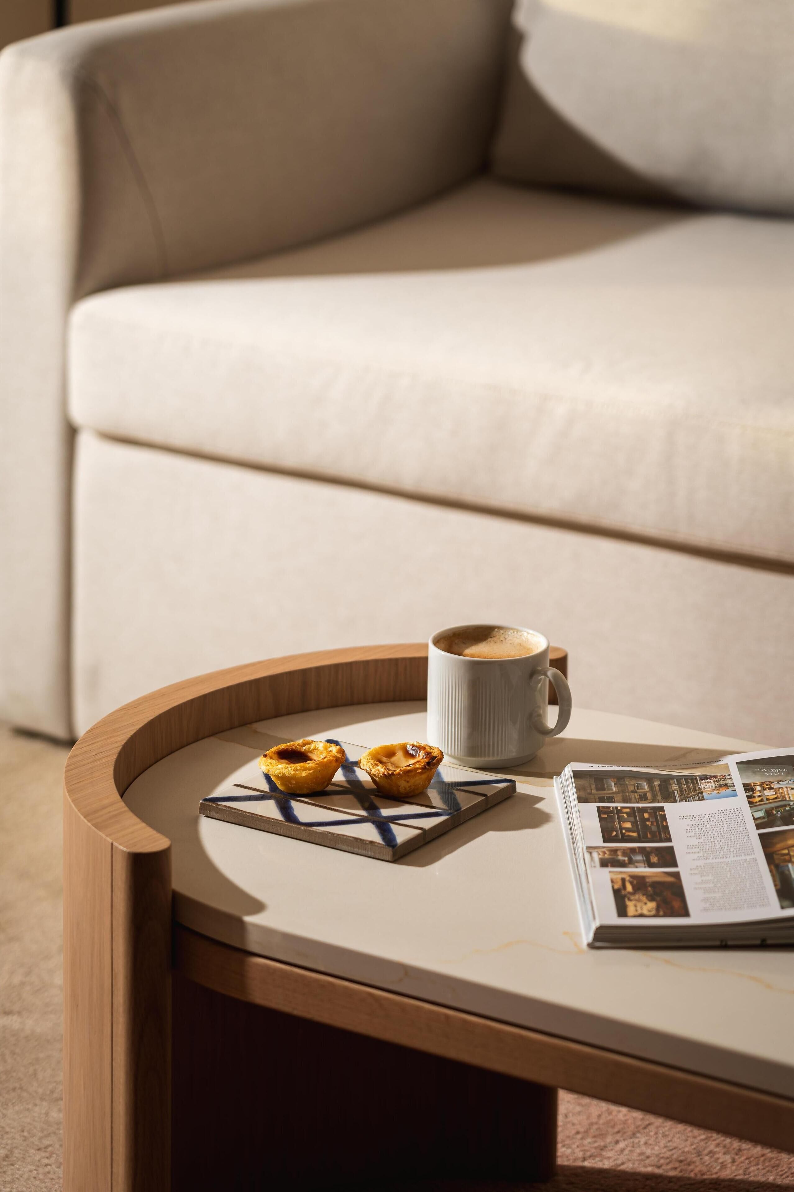A round wooden table with a white surface, a cup of coffee, two pastries, and a magazine.