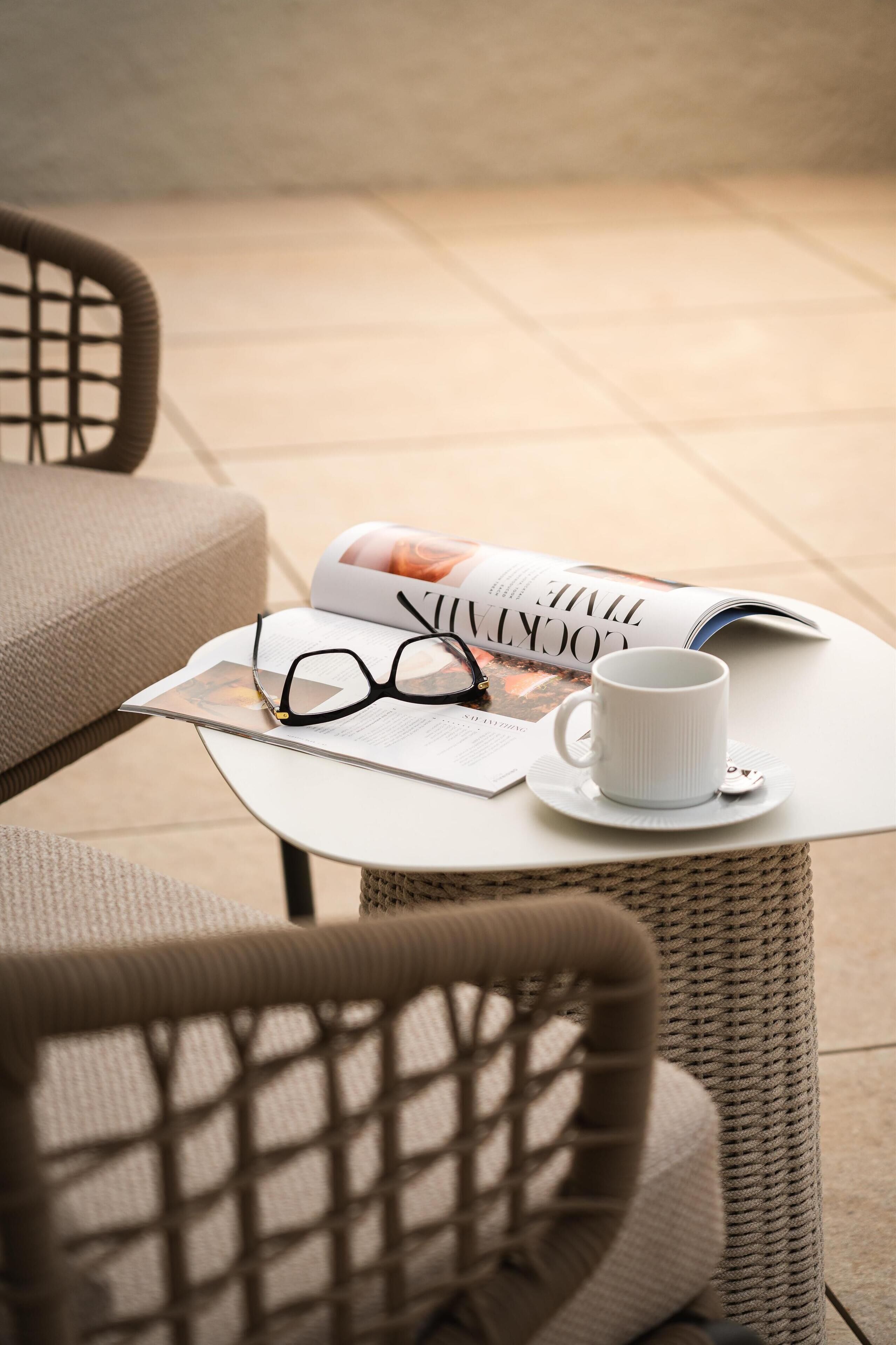 A wicker chair, a white table with a magazine, a pair of glasses, and a white mug.