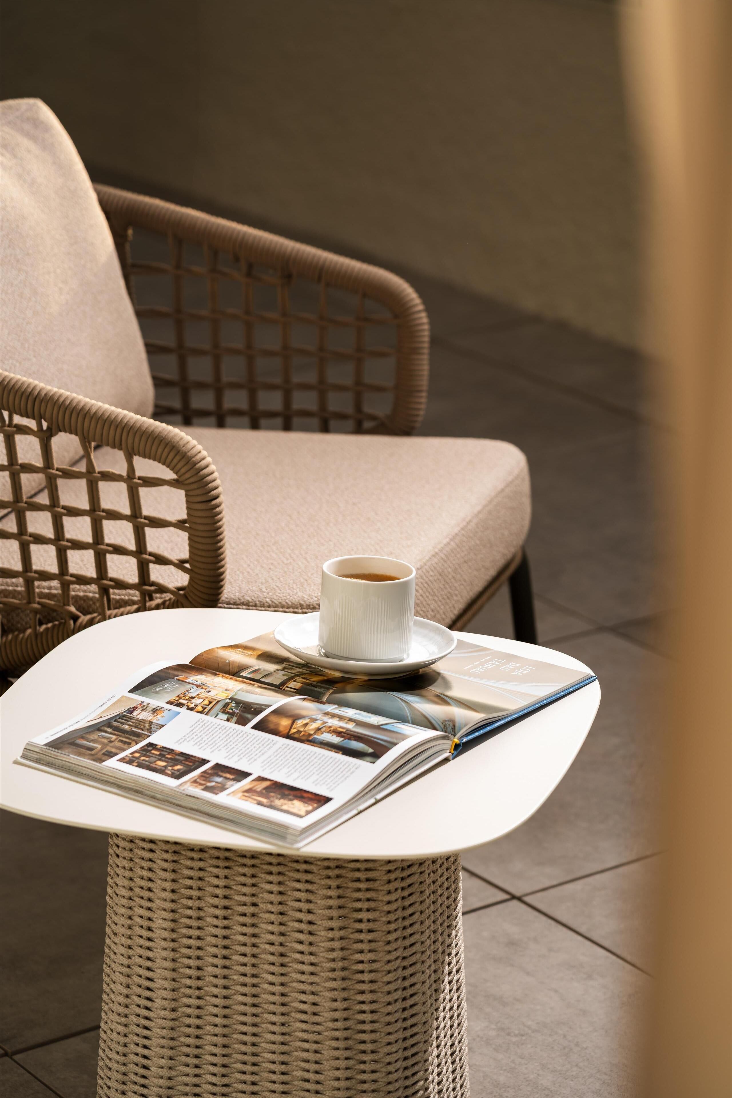 A wicker chair with a cushion, a table with a magazine and a cup of coffee.