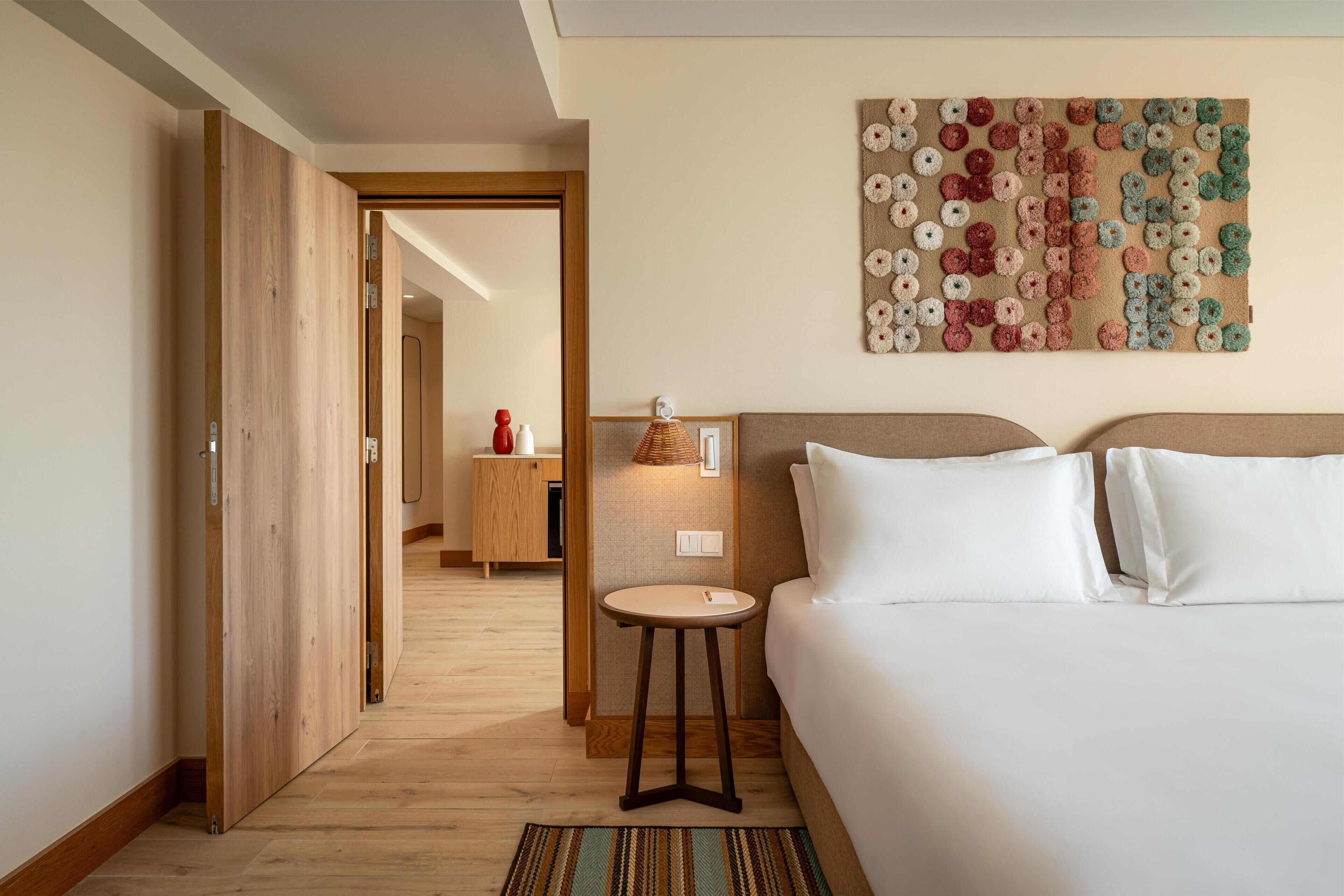 A hotel room with a bed, a small table, a wall art piece, and a wooden door leading to another room.