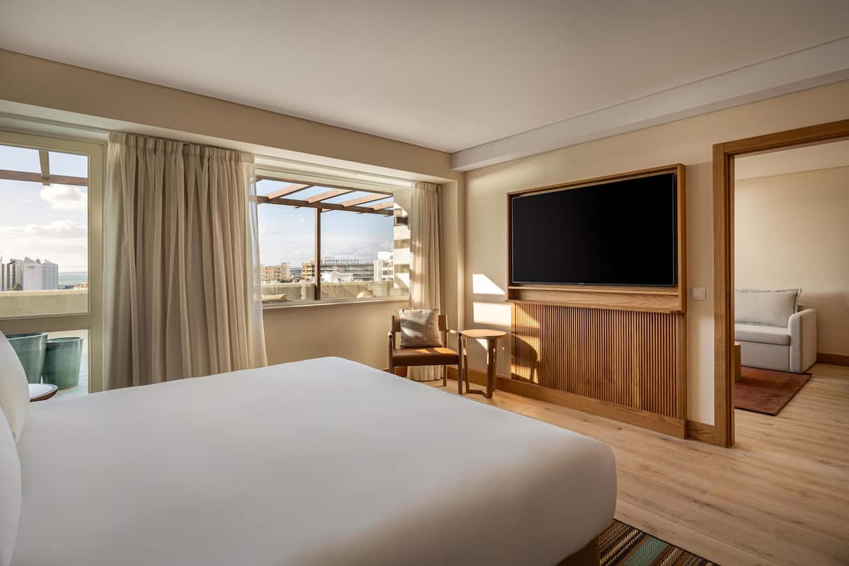 A hotel room with a large bed, a TV, a seating area with a chair and table, and a view of the city.