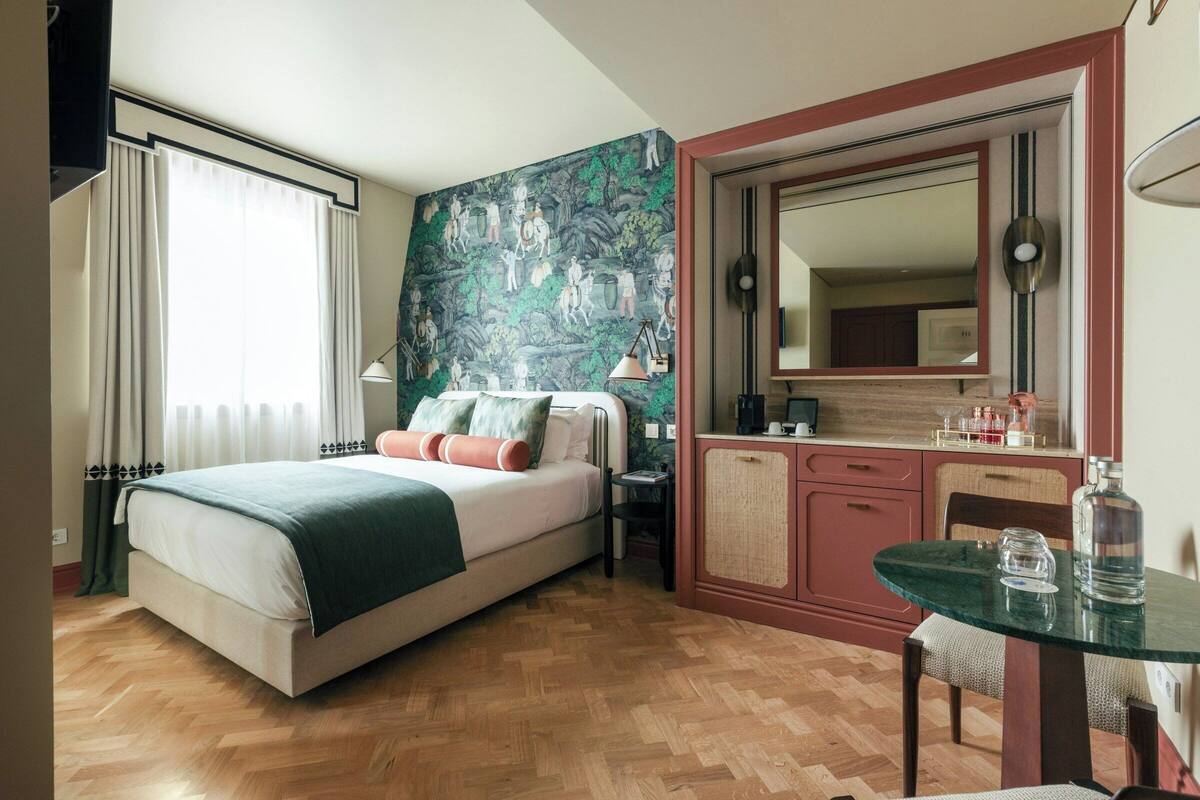 A hotel room with a bed, a desk, a chair, a table, a mirror, and a window with curtains.