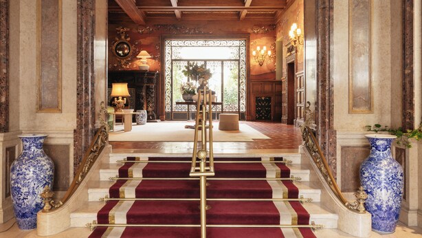 Un magnifique escalier recouvert d’un tapis rouge, bordé de vases bleus, mène à une pièce ornée d’un lustre.