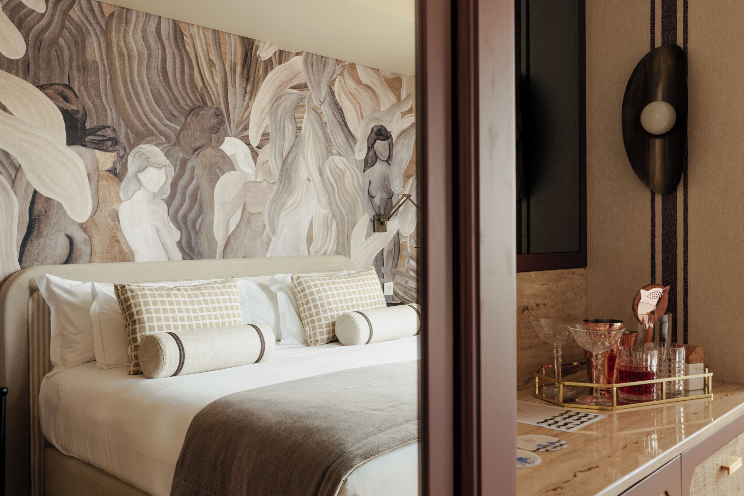 Une chambre à coucher ornée d’une grande fresque murale abstraite, avec un lit recouvert de nombreux coussins, une table de chevet sur laquelle se trouve un plateau avec des verres et une bouteille, et un luminaire fixé au mur.