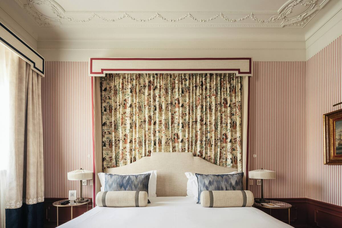 A hotel room with a large bed, striped wallpaper, floral curtains, and two bedside tables with lamps.