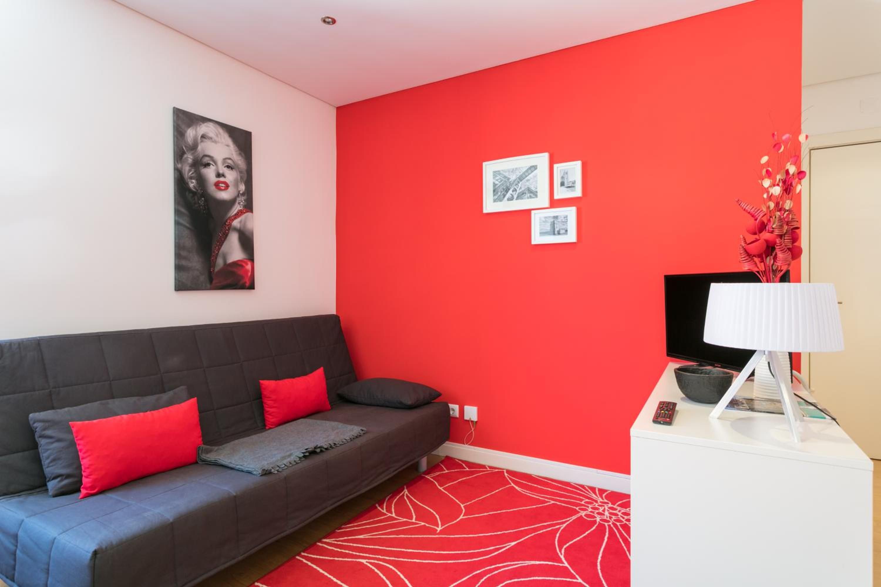 A modern living room with a red accent wall, a grey sofa with red cushions, a white side table with a lamp, and framed pictures.