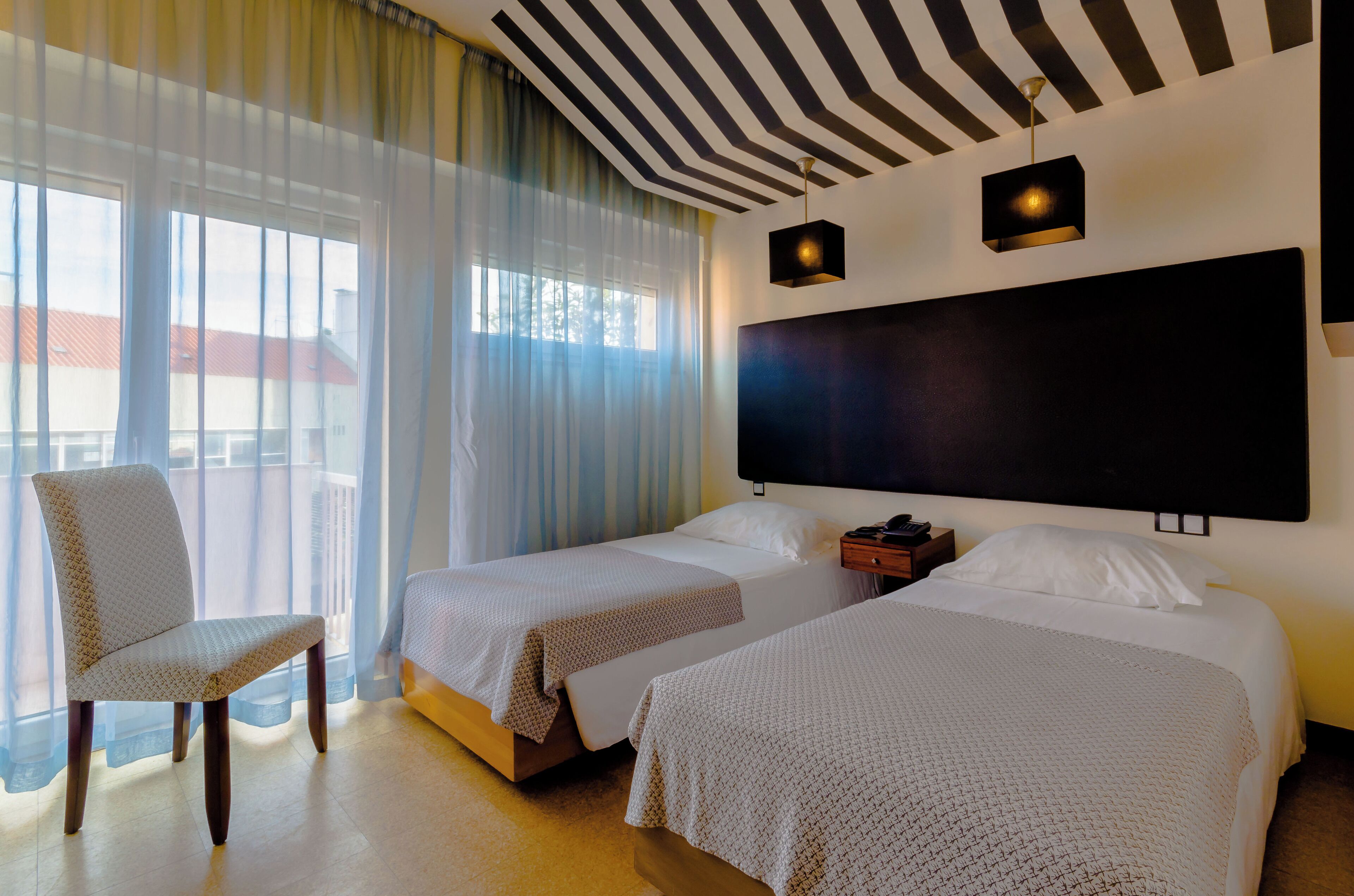 Quarto de hotel com duas camas, uma cadeira, uma janela grande com cortinas, uma cabeceira preta e um lustre.