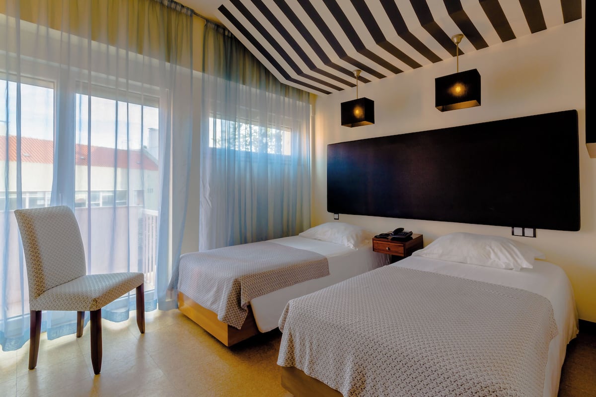 Quarto de hotel com duas camas, uma cadeira, uma janela grande com cortinas, uma cabeceira preta e um lustre.
