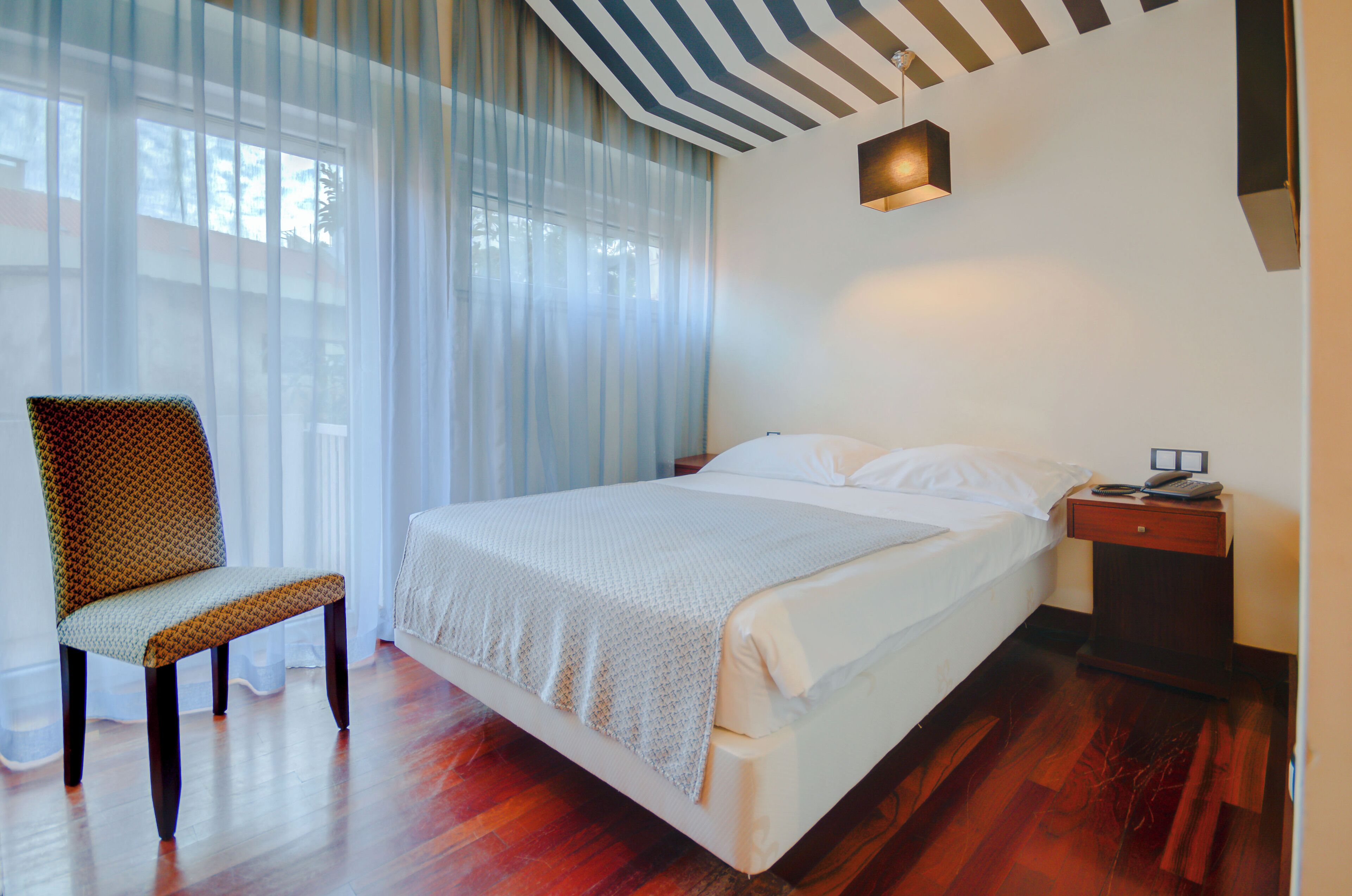 Quarto com cama, cadeira, mesa de cabeceira e janela com cortinas leves.