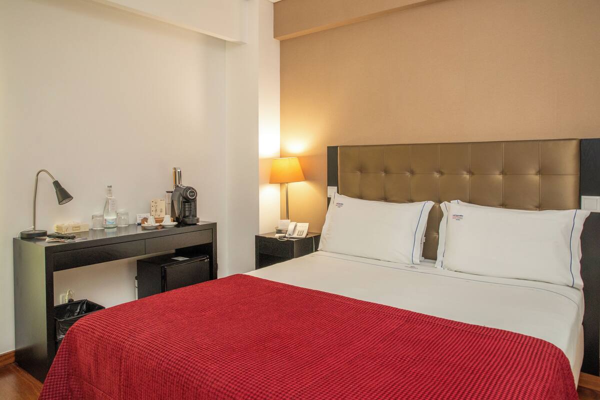 Una camera d'albergo con un letto, una scrivania con una lampada, un telefono e un cestino.