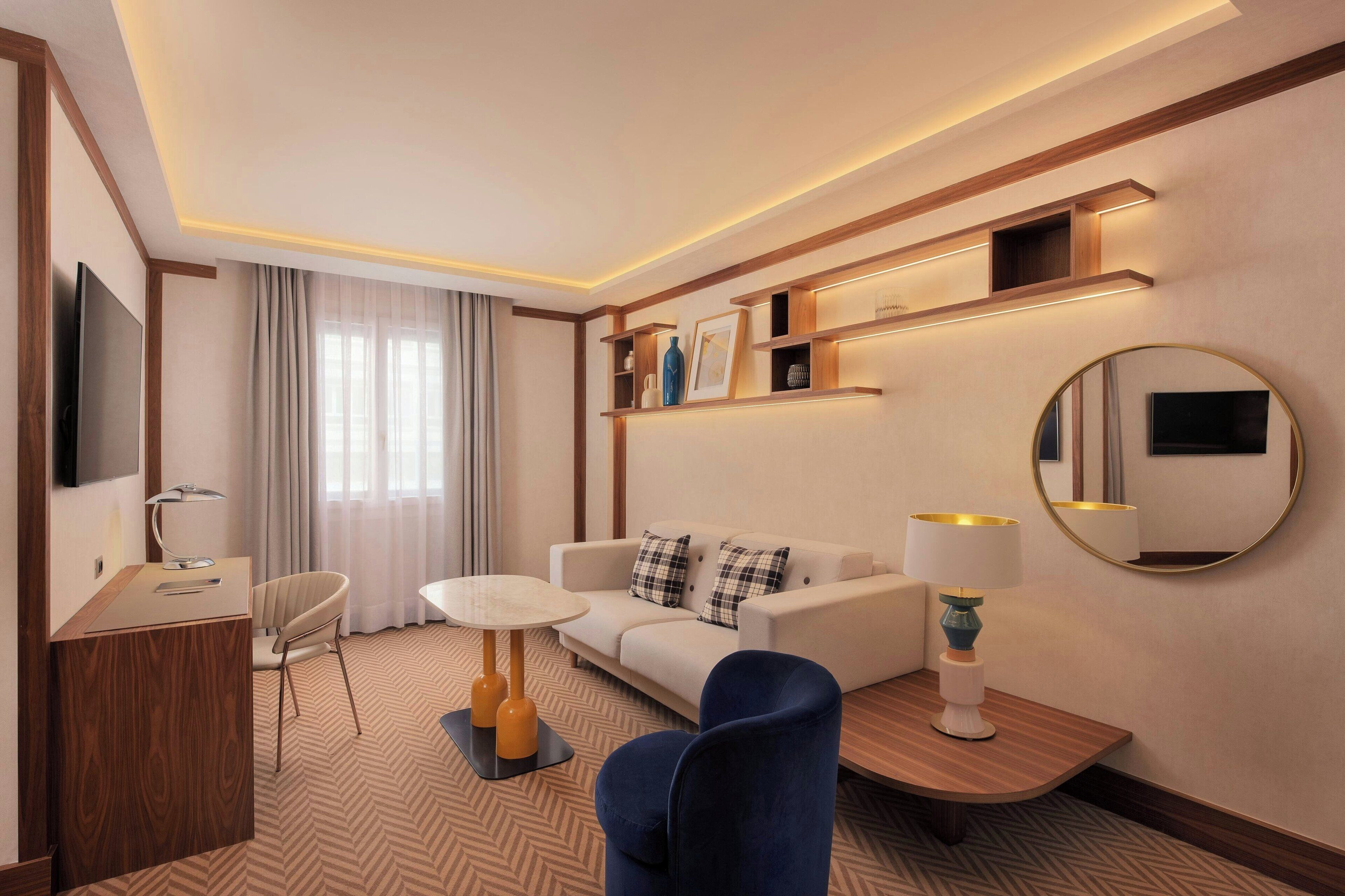 A hotel room with a sofa, a desk, a chair, a round mirror, and shelves with decorative items.