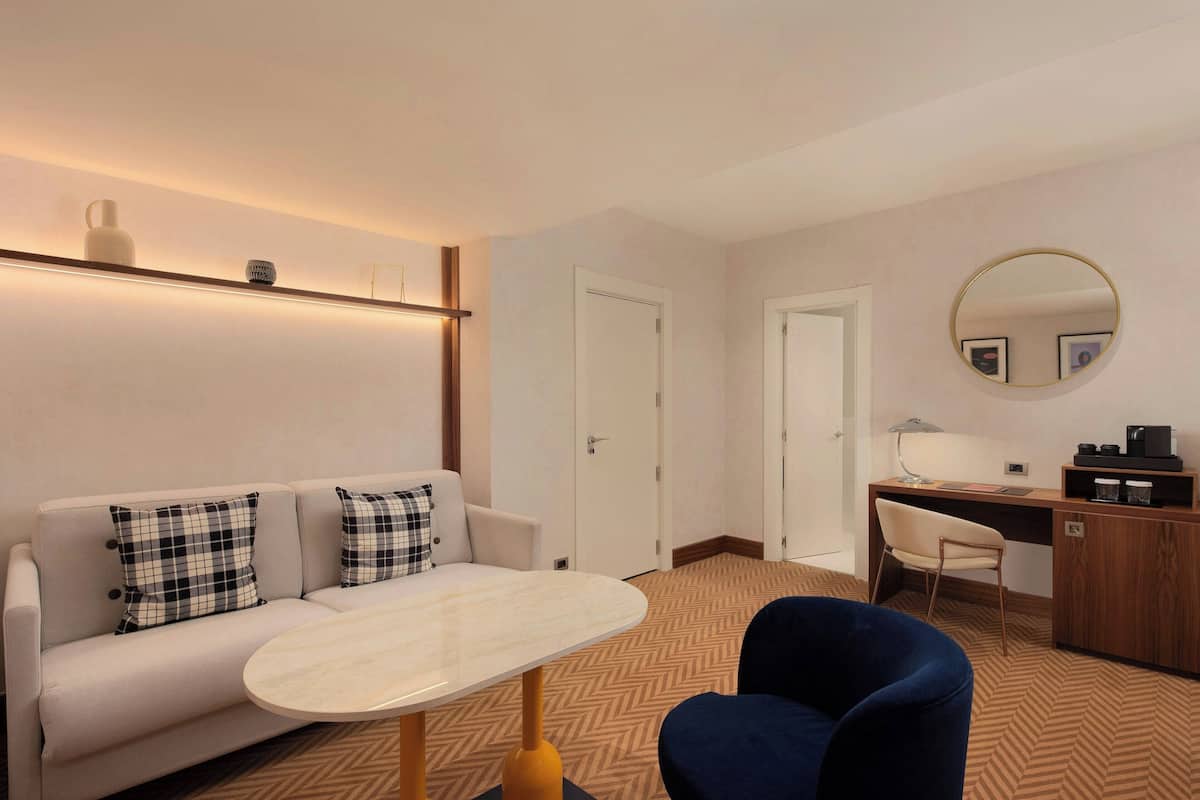 Ein modernes Hotelzimmer mit Sofa, rundem Tisch, Schreibtisch mit Stuhl, Spiegel und Kleiderschrank.