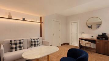 A modern hotel room with a sofa, a round table, a desk with a chair, a mirror, and a wardrobe.