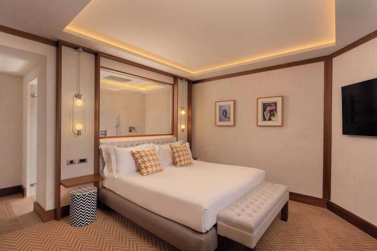 Ein Hotelzimmer mit einem großen Bett, einer Sitzbank, einem Spiegel und einem Fernseher.