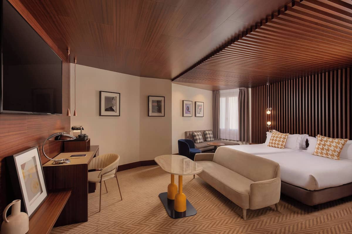 Ein Hotelzimmer mit Holzboden, einem Bett mit gemusterten Kissen, einer Couch, einem Schreibtisch mit Lampe und gerahmten Bildern an der Wand.