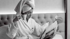 Une personne en peignoir, assise sur un lit, avec un journal, une tasse et un plateau sur lequel se trouvent une théière et des tasses.