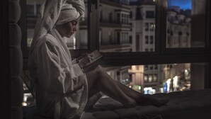 Une personne vêtue d’une robe, assise près d’une fenêtre, avec une vue sur la ville.