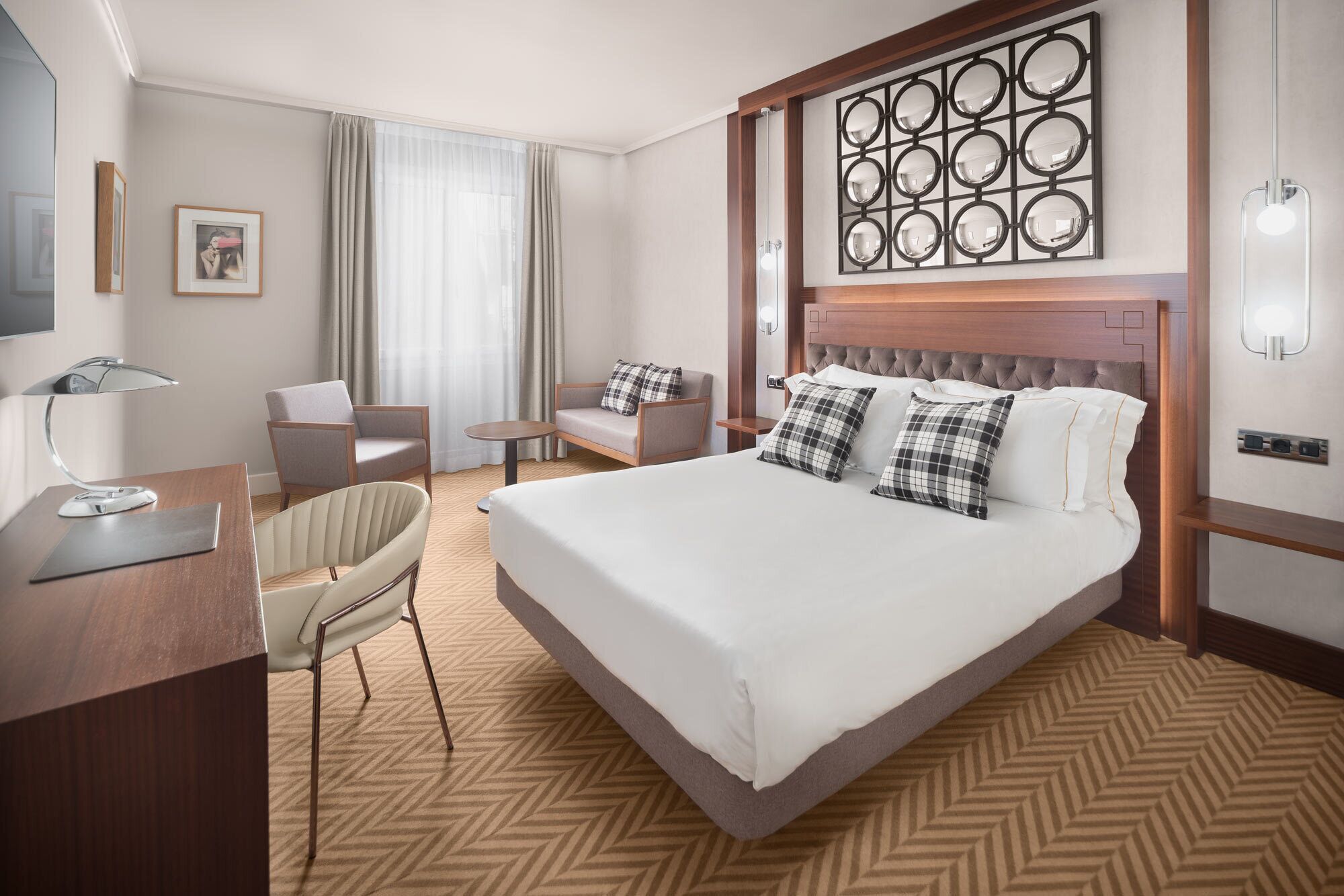 A hotel room with a large bed, a desk with a lamp, two armchairs, a small table, and a decorative wall panel.