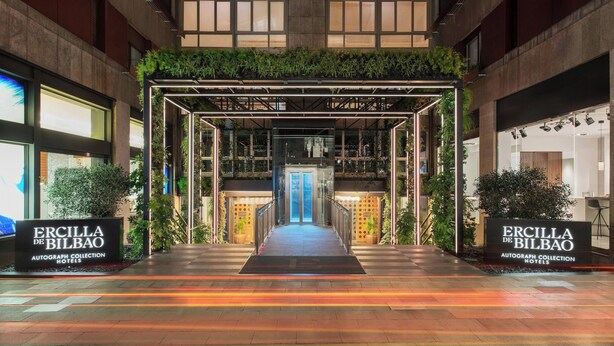 L’entrée de l’hôtel ERCILLA DE BILBAO AUTOGRAPH COLLECTION se distingue par sa façade en verre, sa porte centrale bleue et son auvent métallique soutenu par de la verdure.