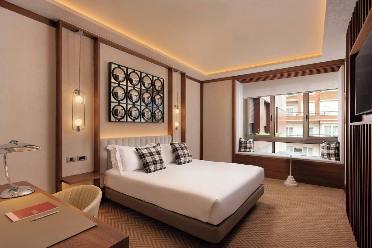 Ein Hotelzimmer mit einem großen Bett, einem Schreibtisch mit Lampe, einem wandmontierten Spiegel und einem Fenster mit Blick auf Gebäude.