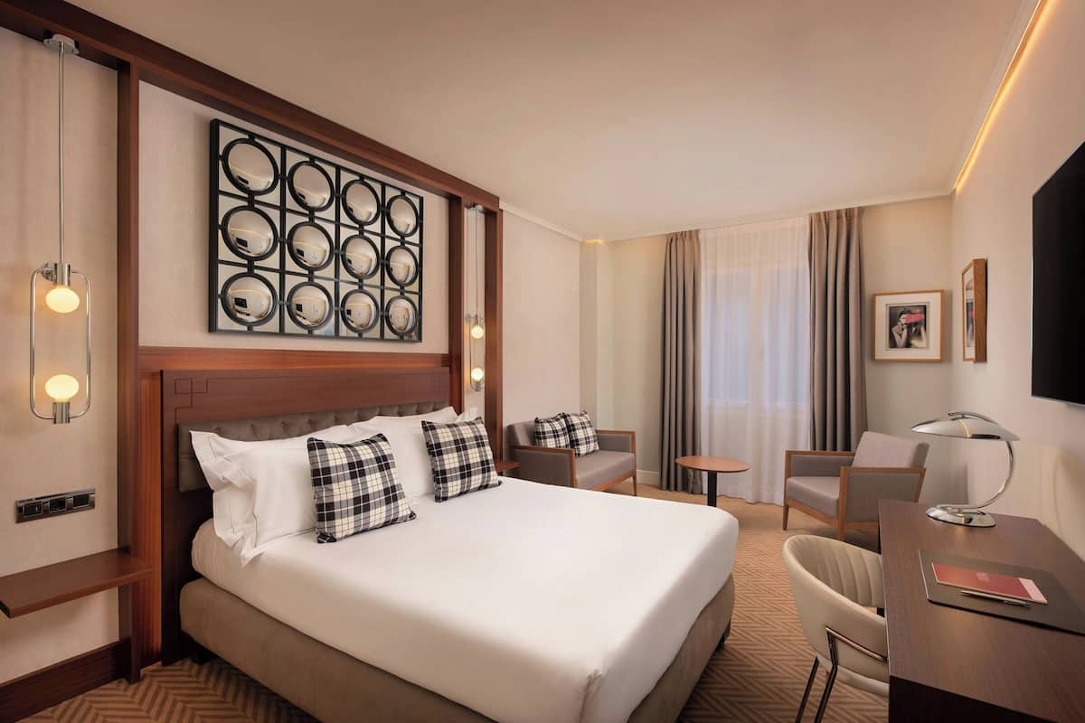 Ein Hotelzimmer mit einem großen Bett, zwei Sesseln, einem Schreibtisch, einem Wandspiegel und einem Fernseher.