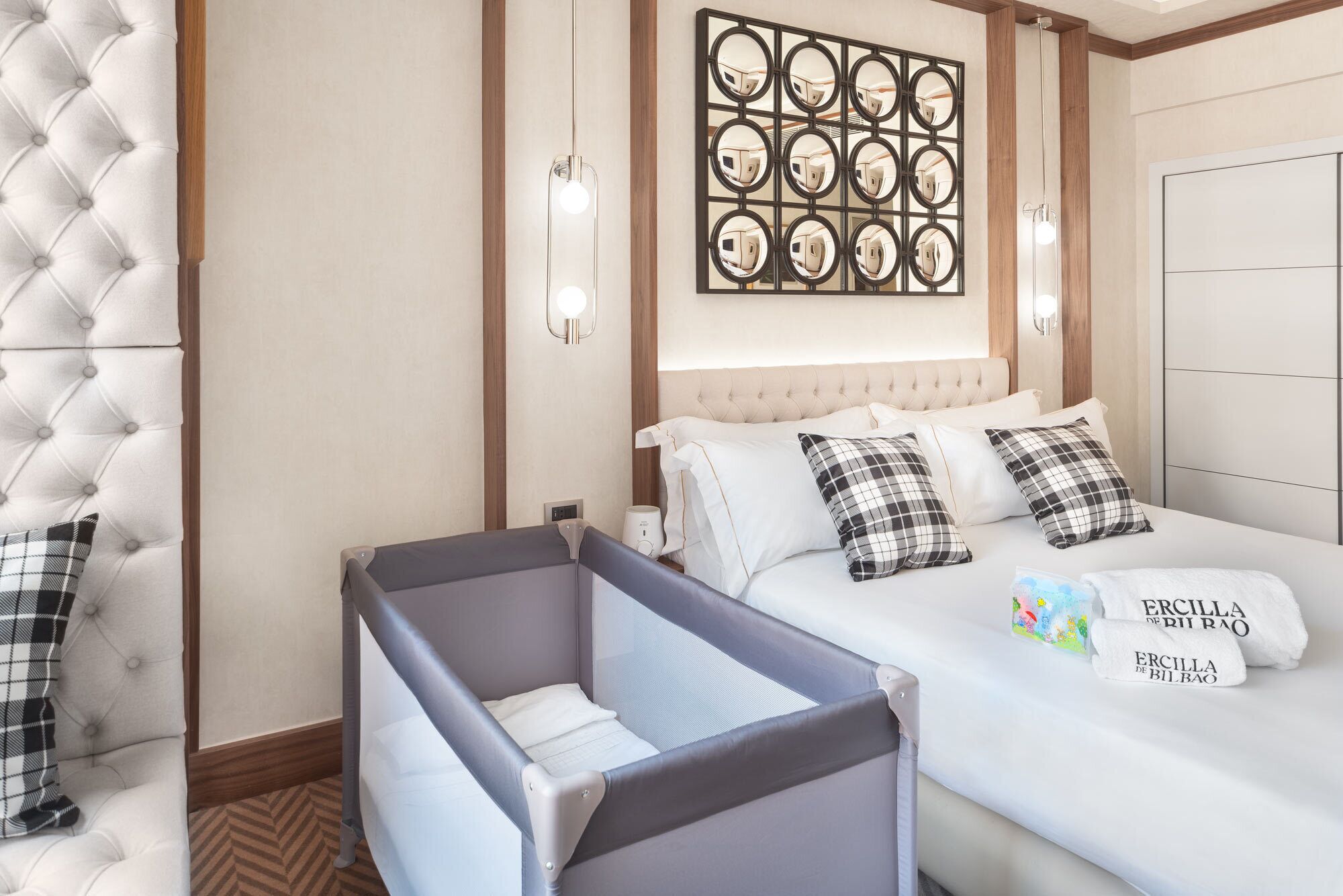 Habitación de hotel con una cama grande, una cuna para bebé, un adorno de pared y una mesita de noche con lámpara.