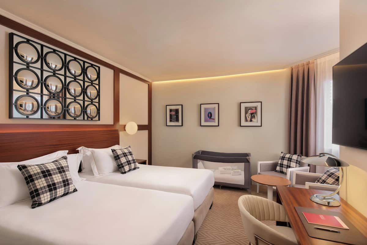 Ein Hotelzimmer mit zwei Betten, einem Schreibtisch mit Stuhl, einem Fernseher und einem Wandspiegel.