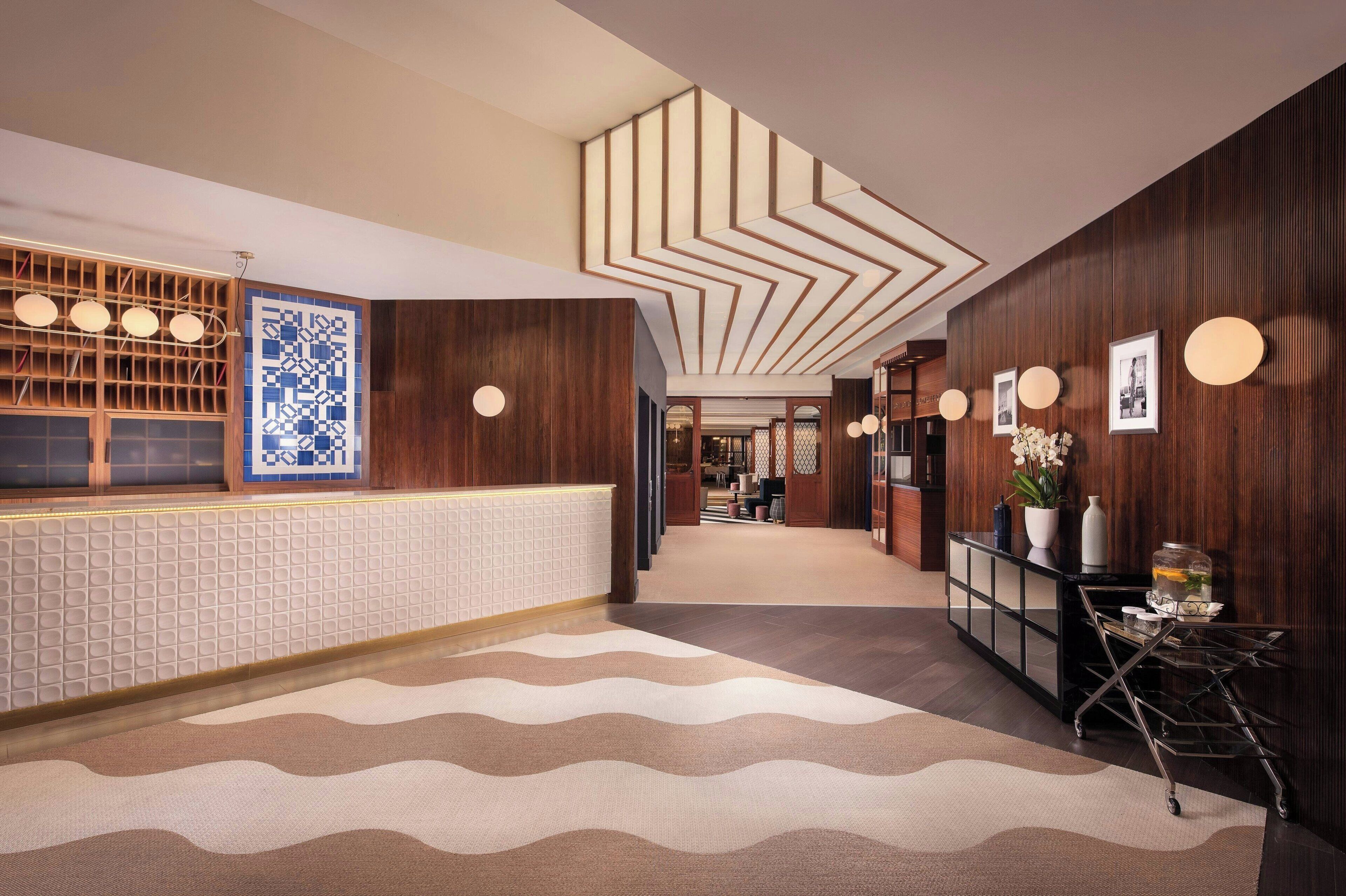 Vestíbulo de hotel con mostrador de recepción, pared con diseño, techo moderno y un carro decorativo.