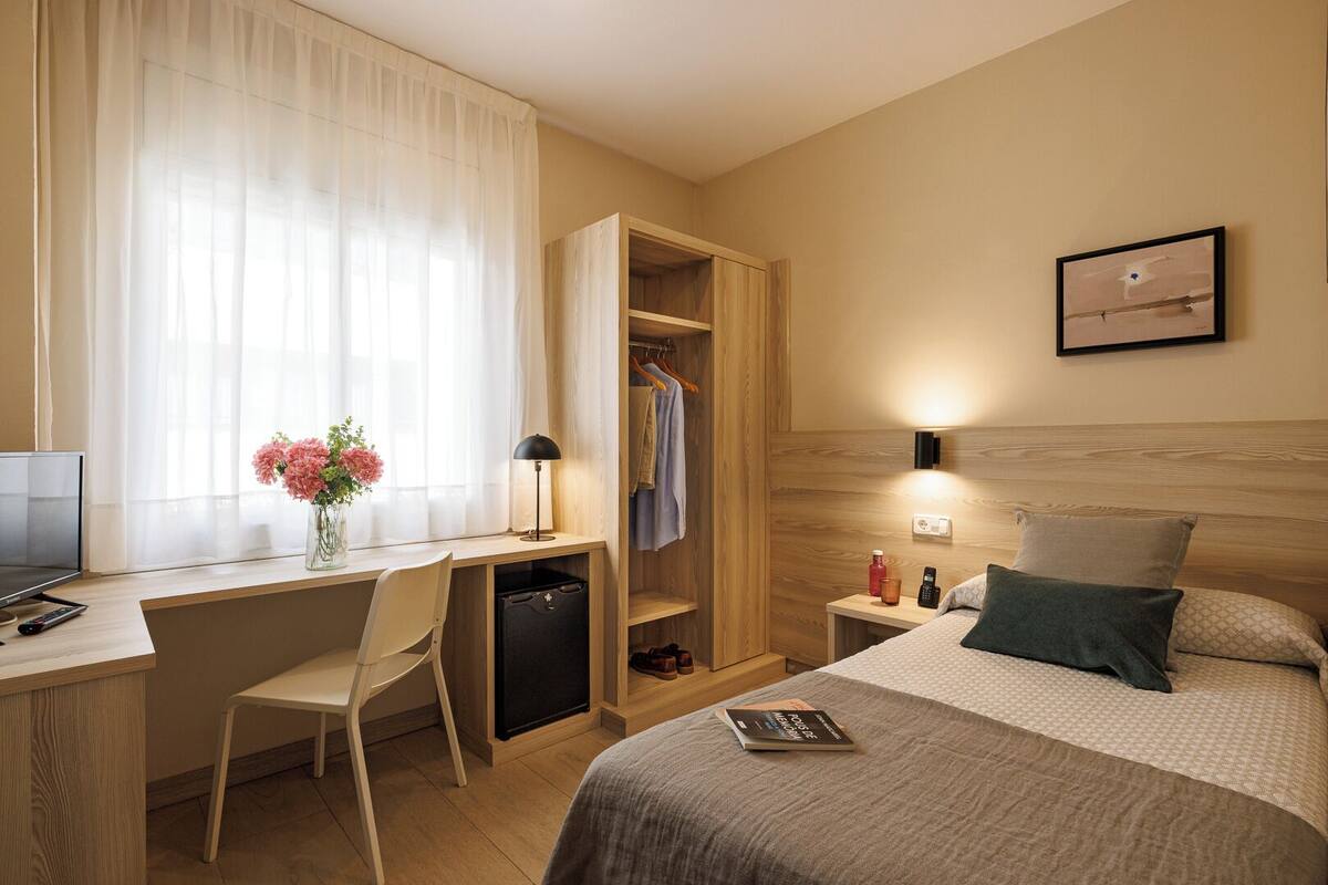 A hotel room with a desk, chair, bed, bedside table, lamp, and a wardrobe.