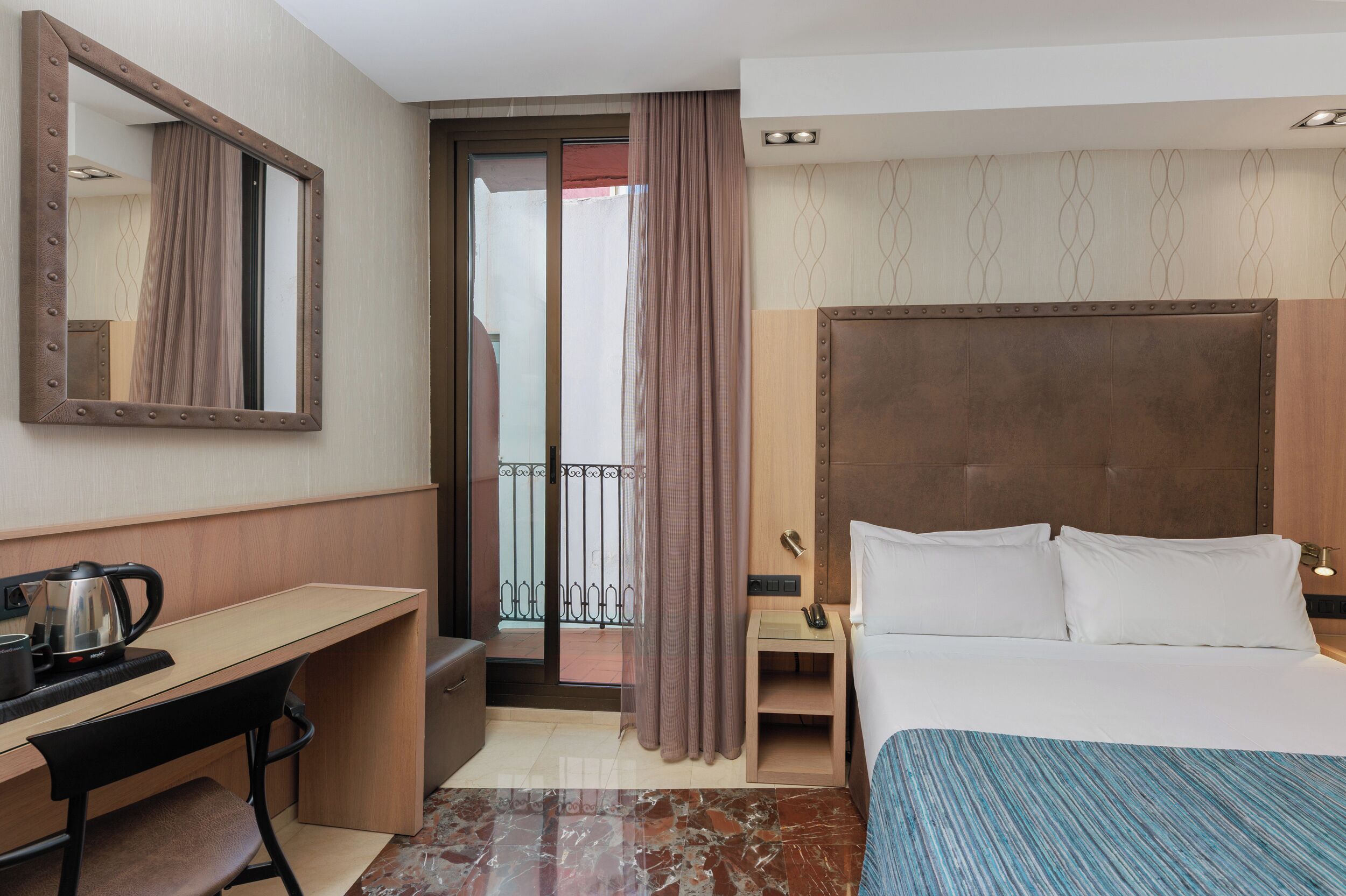 A hotel room with a bed, a desk with a kettle, a mirror, a small table with a phone, and a view of the outside through a door.