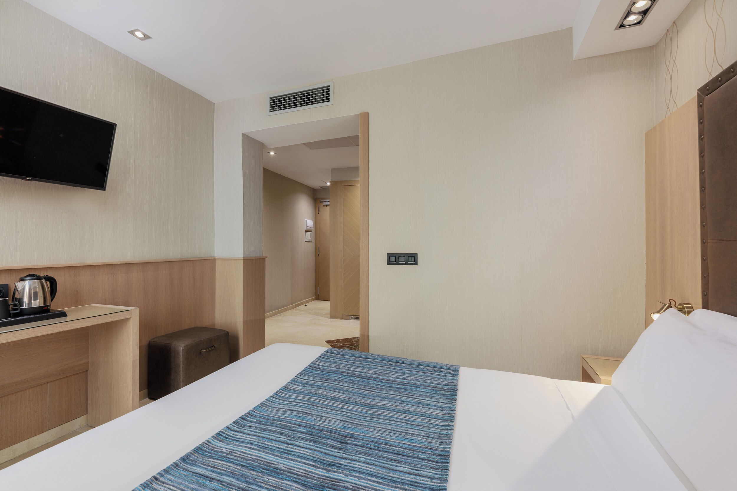A hotel room with a bed, a flat-screen TV mounted on the wall, a desk with a kettle, and a suitcase.
