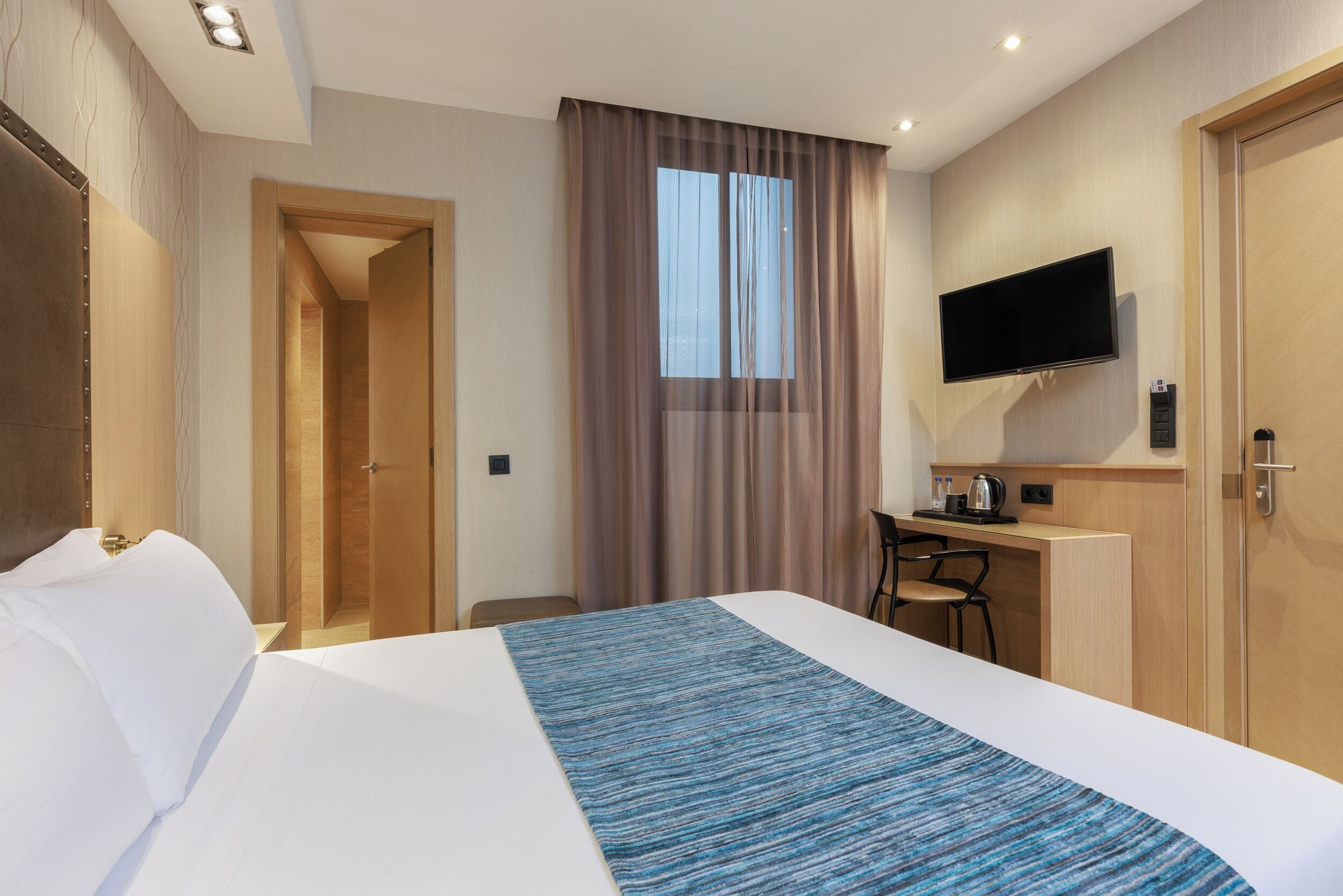 Habitación de hotel con cama, escritorio, silla, televisor y ventana con cortinas.
