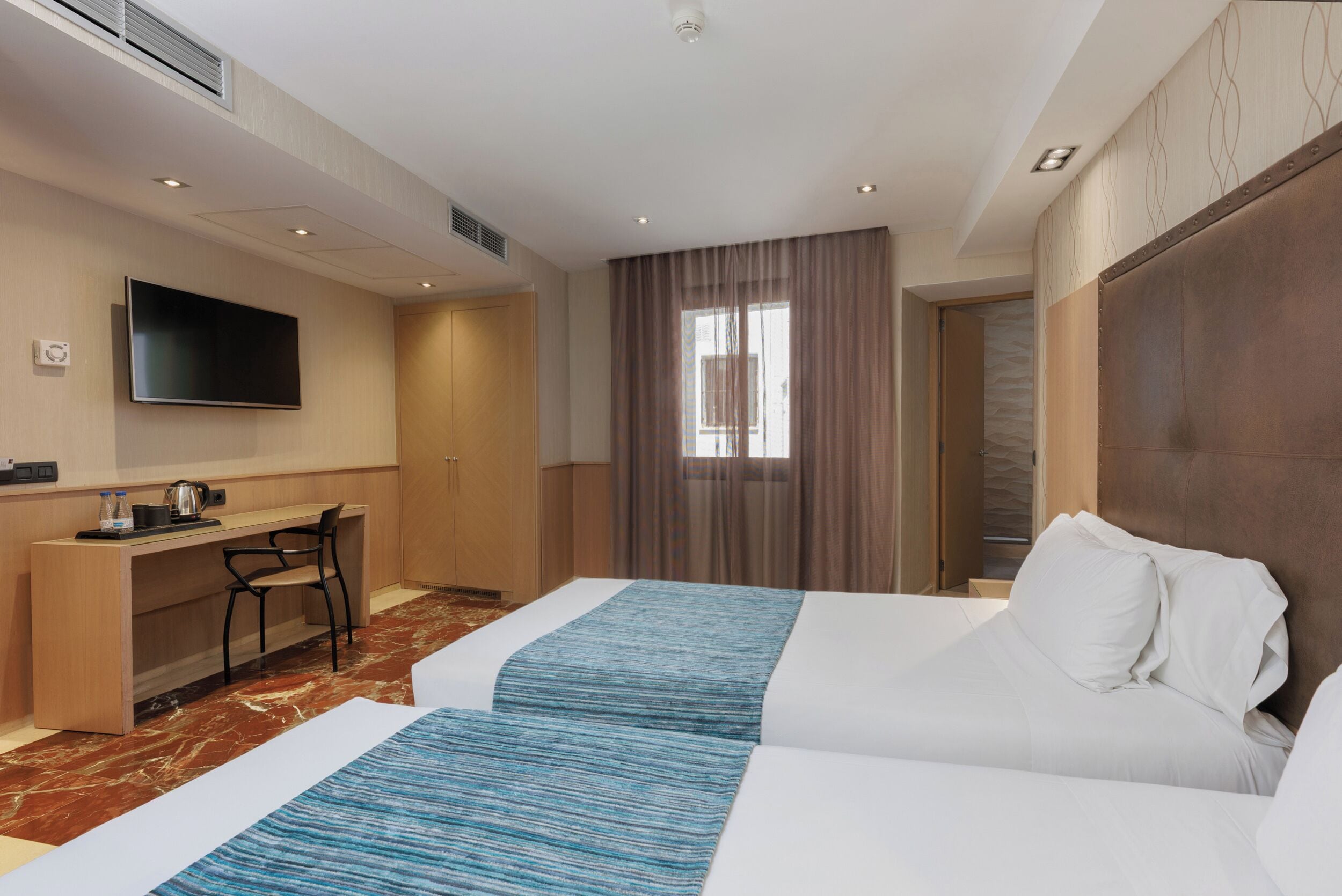 A hotel room with two beds, a desk, a flat-screen TV, and a large window with curtains.