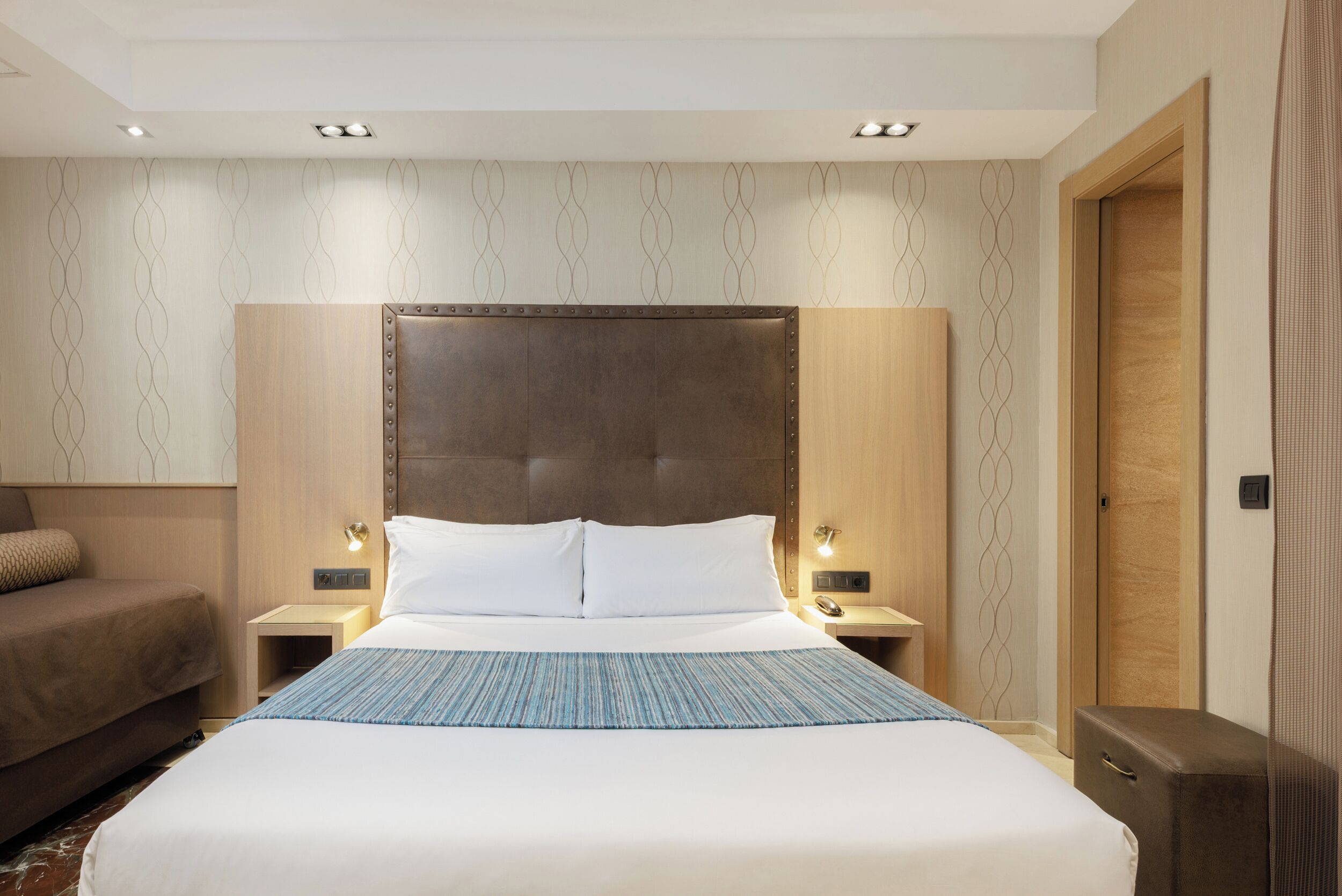 Habitación de hotel con una cama grande, dos mesitas de noche, una lámpara de pared y una puerta.