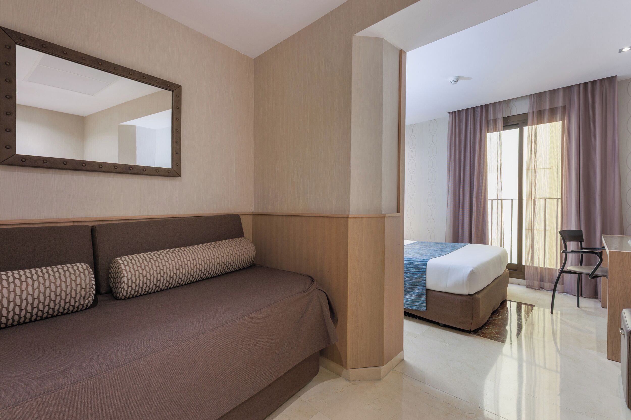 Una habitación de hotel moderna con una cama grande, un sofá con cojines estampados, un escritorio y un espejo grande en la pared.