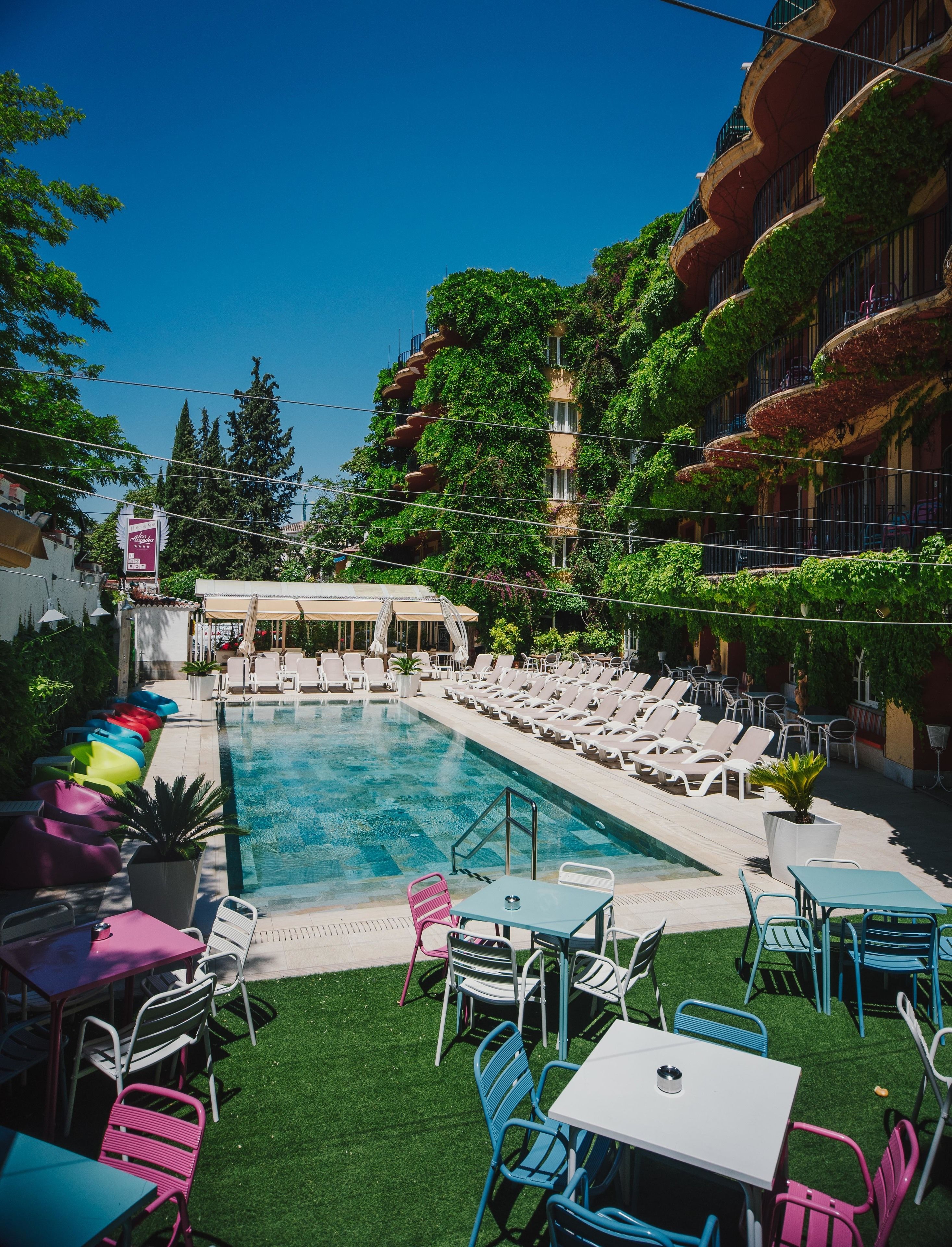 Un área de piscina con sillones y mesas, rodeada de vegetación y un edificio con balcones.