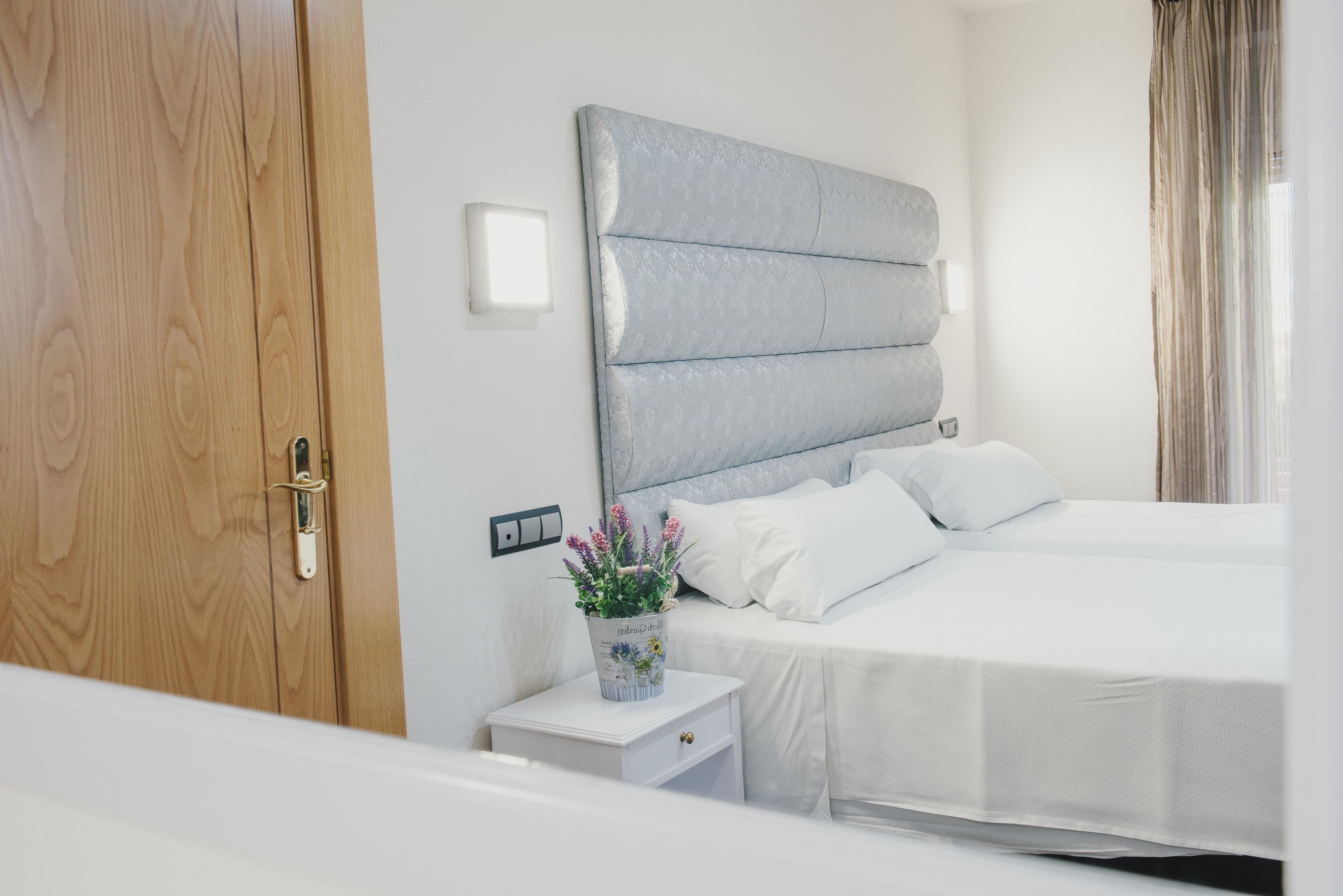 A hotel room with a wooden door, a white bedside table with a potted plant, a bed with white bedding, and a headboard.