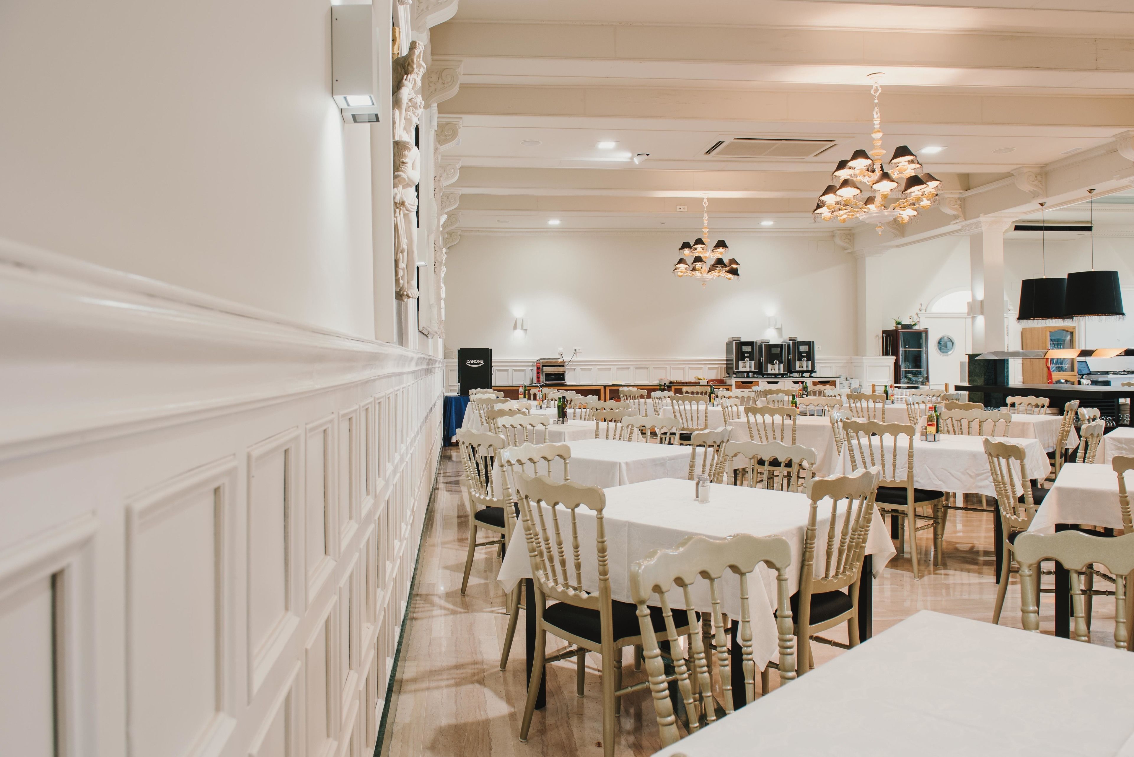 A spacious dining area with multiple tables and chairs, white walls, and a chandelier.