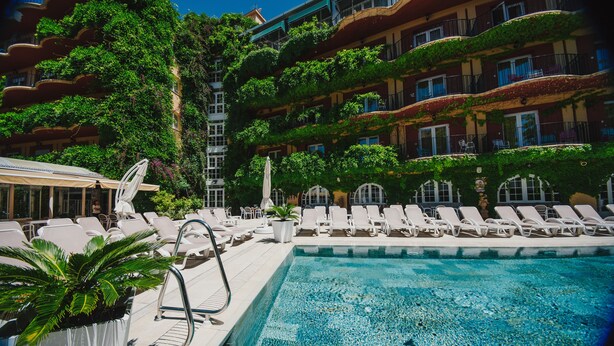 Zona de piscina con sillones de descanso y sombrillas, rodeada de abundante vegetación y un edificio de varios pisos con balcones.
