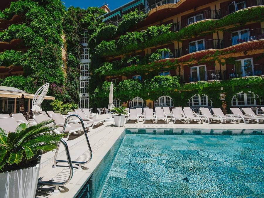 Área de piscina com espreguiçadeiras e guarda-sóis, cercada por vegetação exuberante e um edifício com varandas.