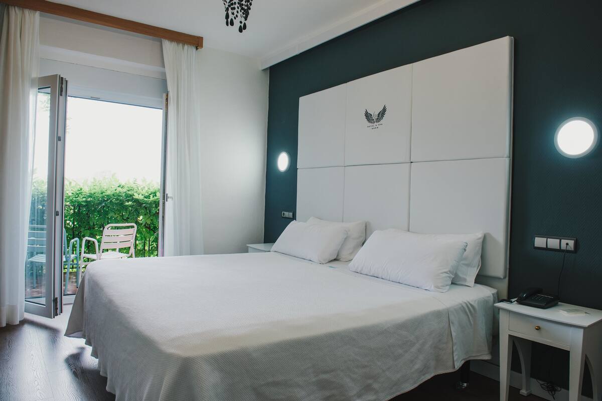 A hotel room with a large bed, a nightstand, a chair by the window, and a view of greenery outside.