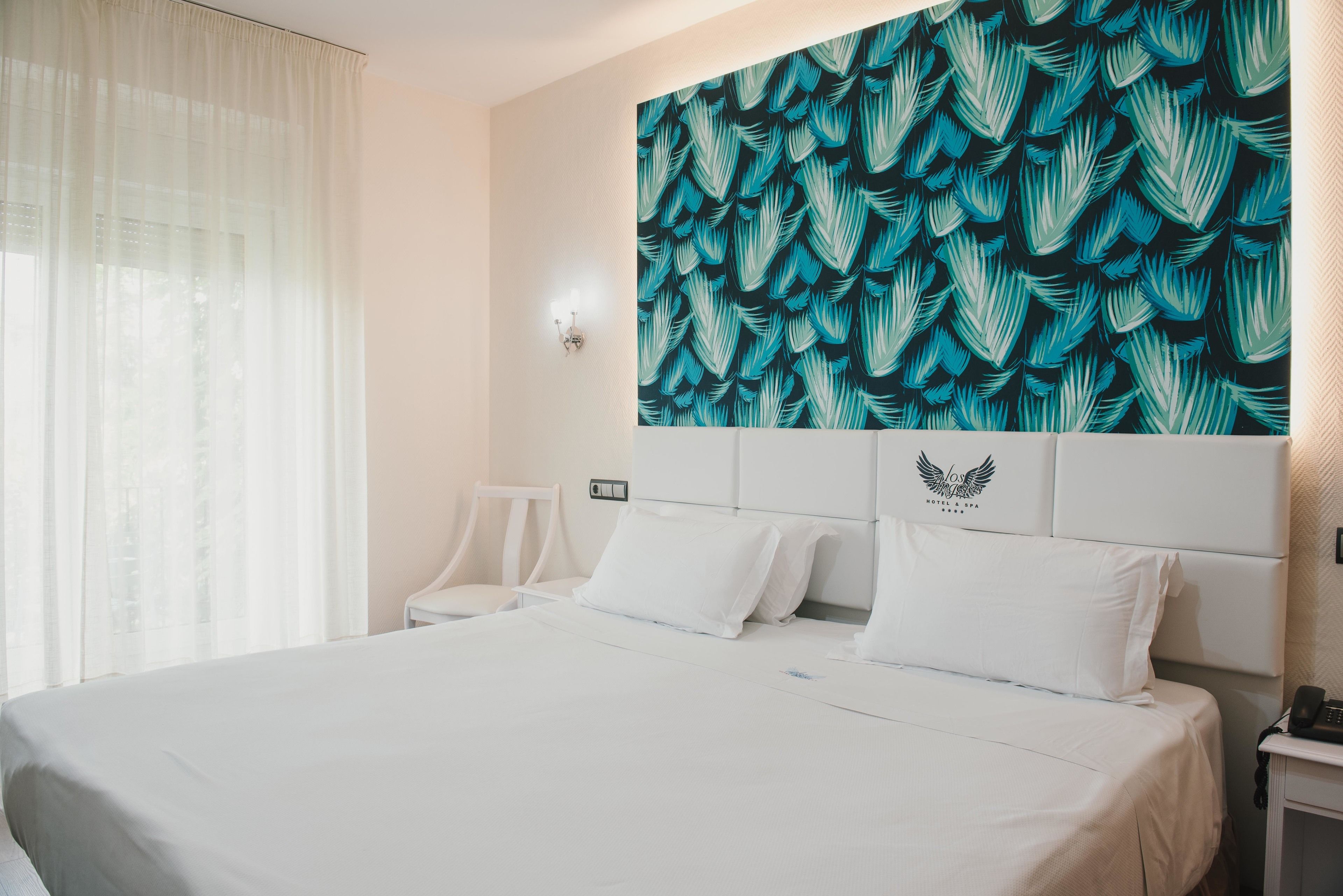 Habitación de hotel con una cama grande, cabecera blanca, pared con diseño de hojas, una silla y una mesita de noche con un teléfono.