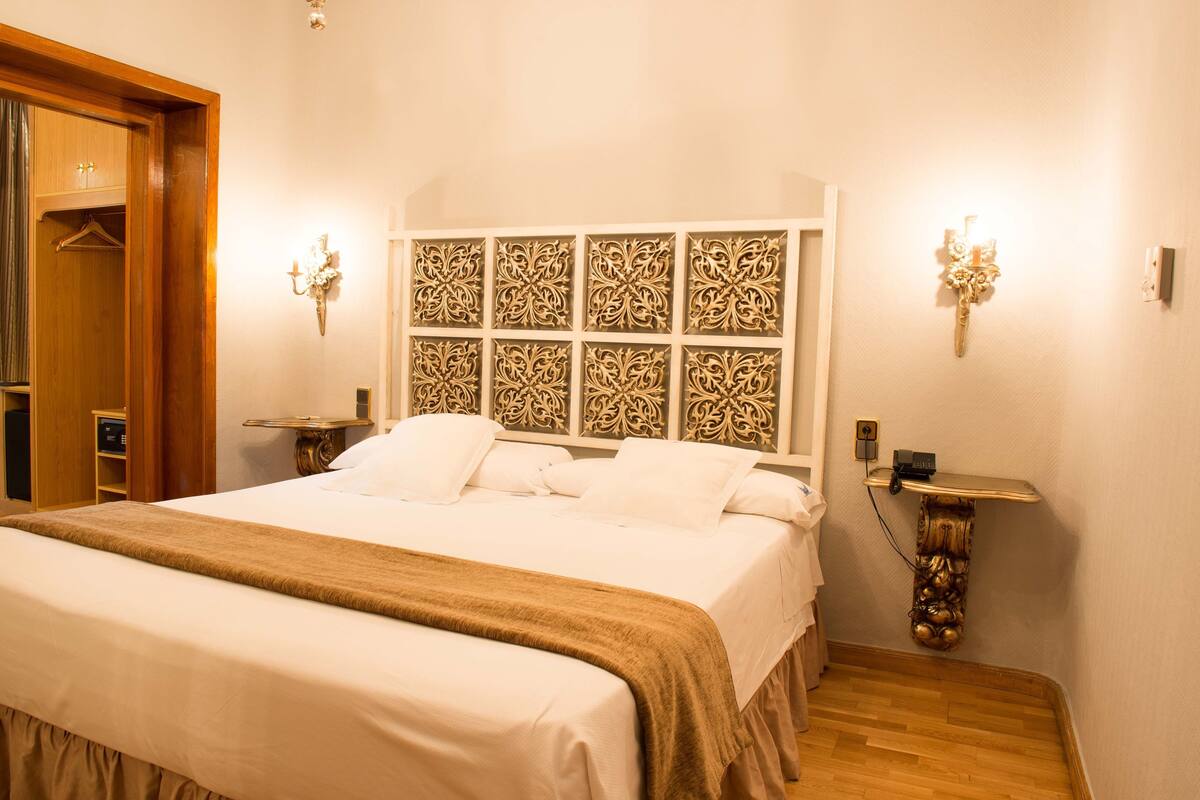 A neatly made bed with a patterned headboard, a bedside table with a lamp, and a wooden floor.