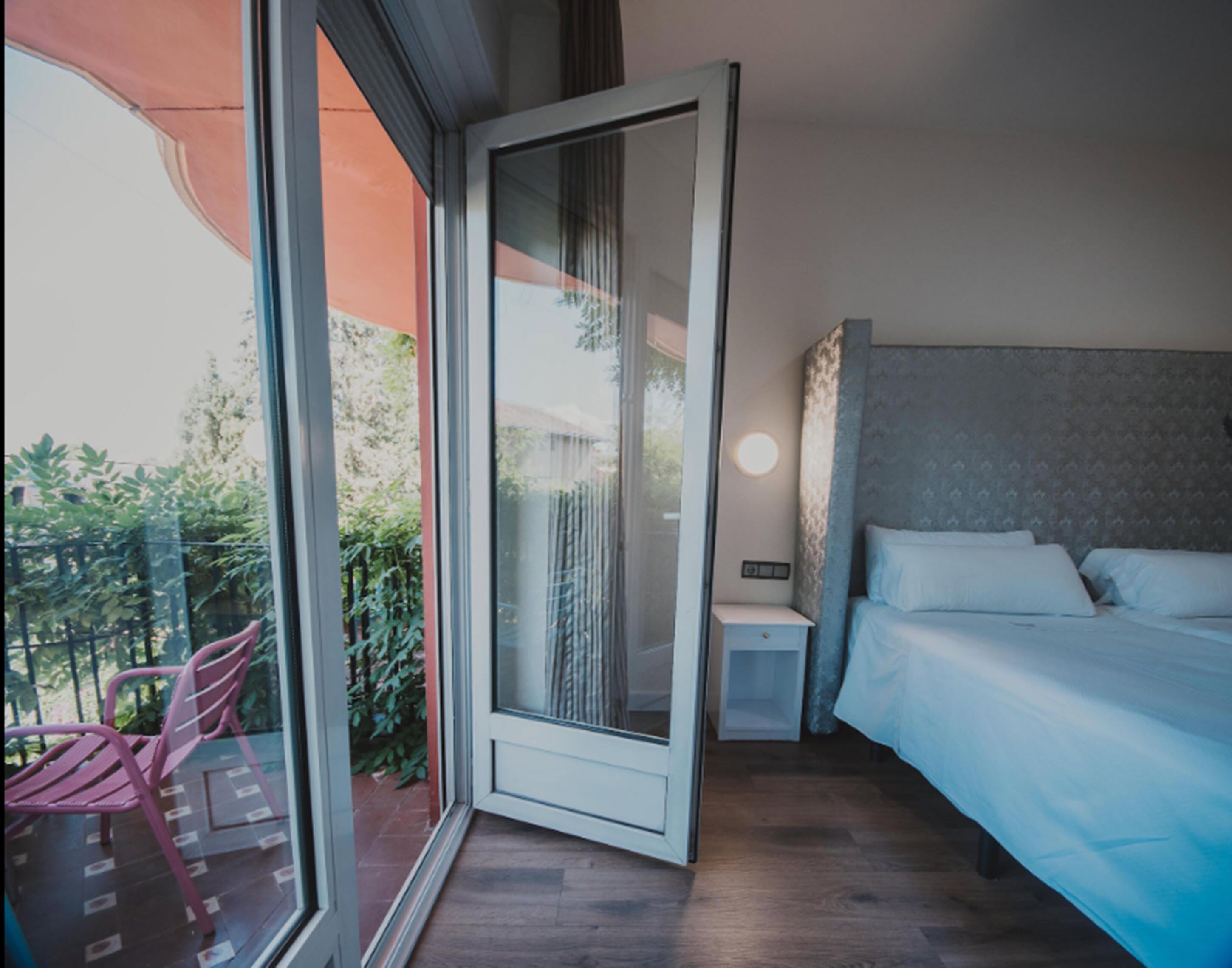 Un dormitorio moderno con un amplio ventanal corredera que da a un balcón, una cama bien hecha y una mesita de noche pequeña.