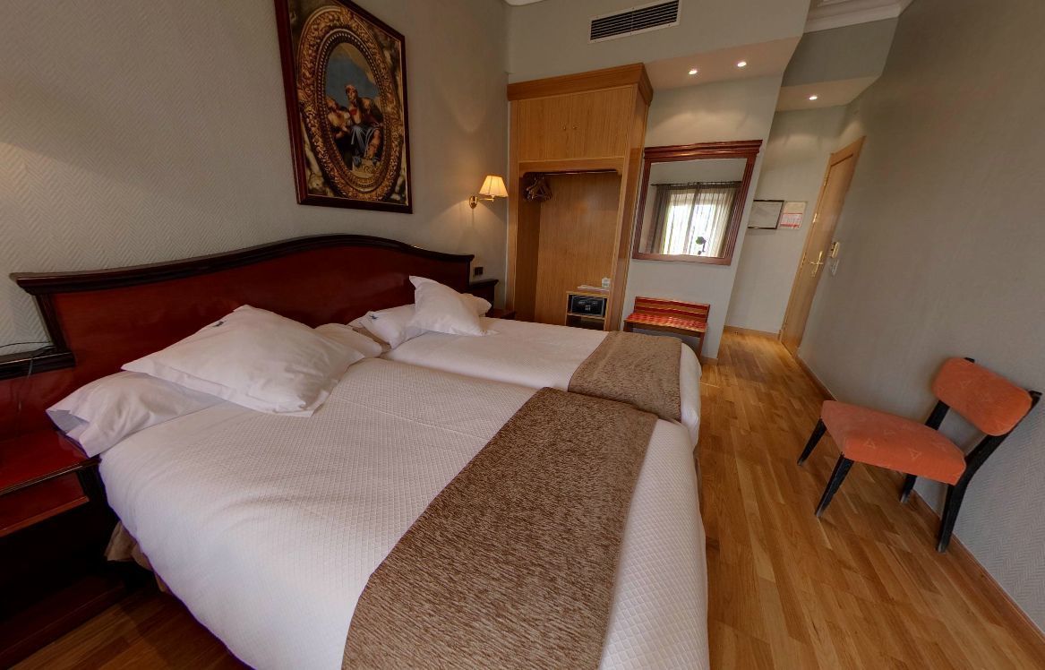 Habitación de hotel con una cama grande, cabecera de madera, un cuadro en la pared, un espejo y un rincón de descanso con dos sillones.
