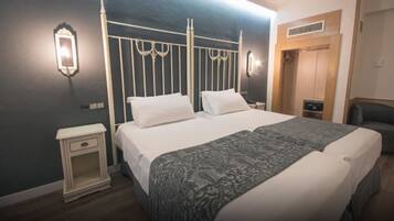 Una cama doble con cabecera blanca y dos mesitas de noche, una manta gris estampada y un armario de madera en una habitación de hotel.