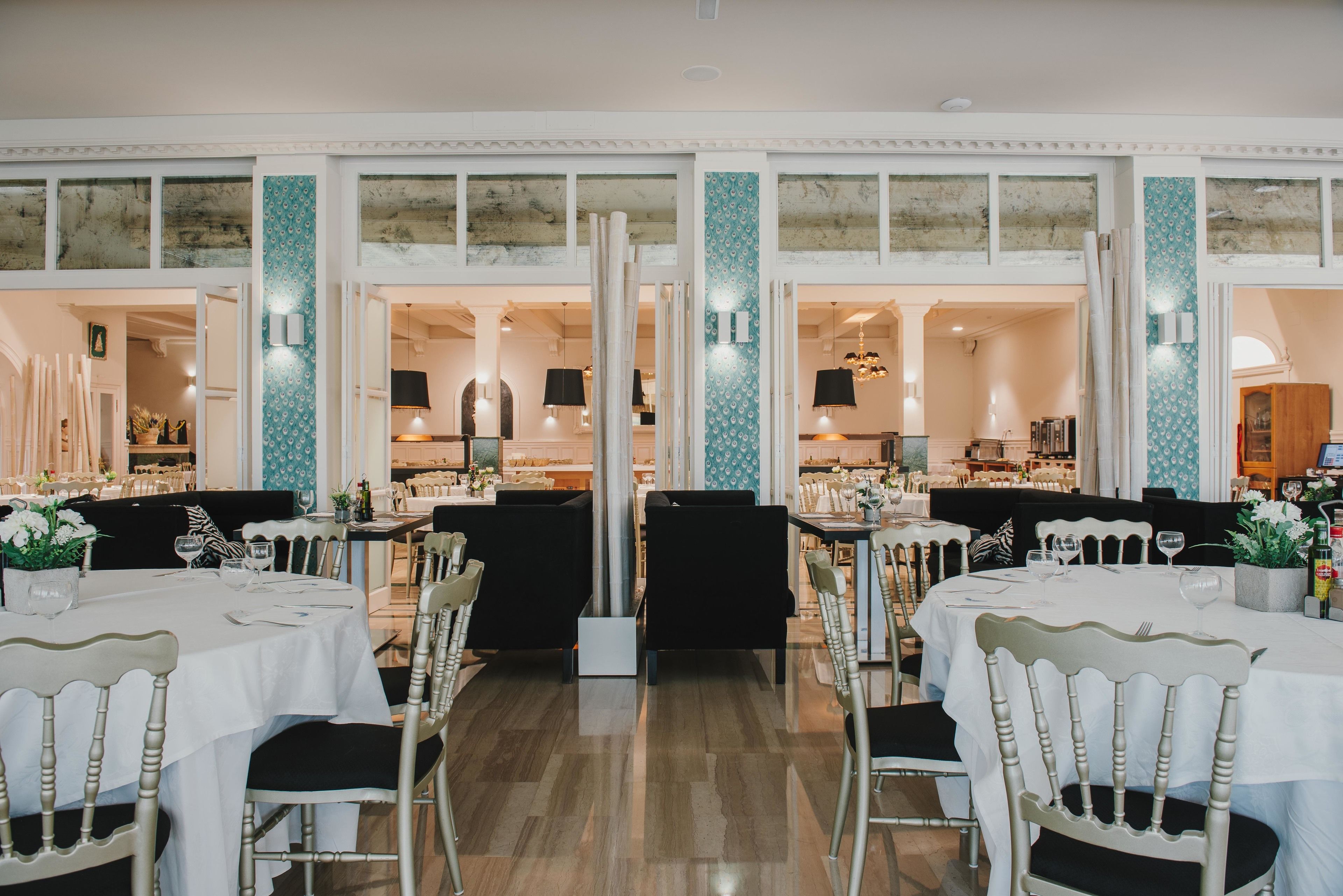 Un restaurante con varias mesas y sillas, piso de madera y vista a la cocina a través de puertas abiertas.