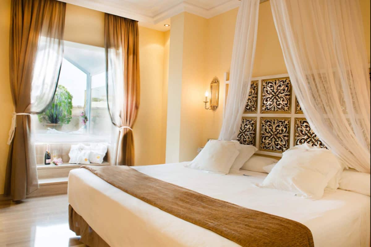 A hotel room with a large bed, a window with curtains, a decorative headboard, and a bedside table with a bottle and glasses.