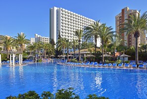 A large swimming pool with blue lounge chairs, palm trees, and a tall hotel building in the background. - Sol Pelícanos Ocas (Benidorm)